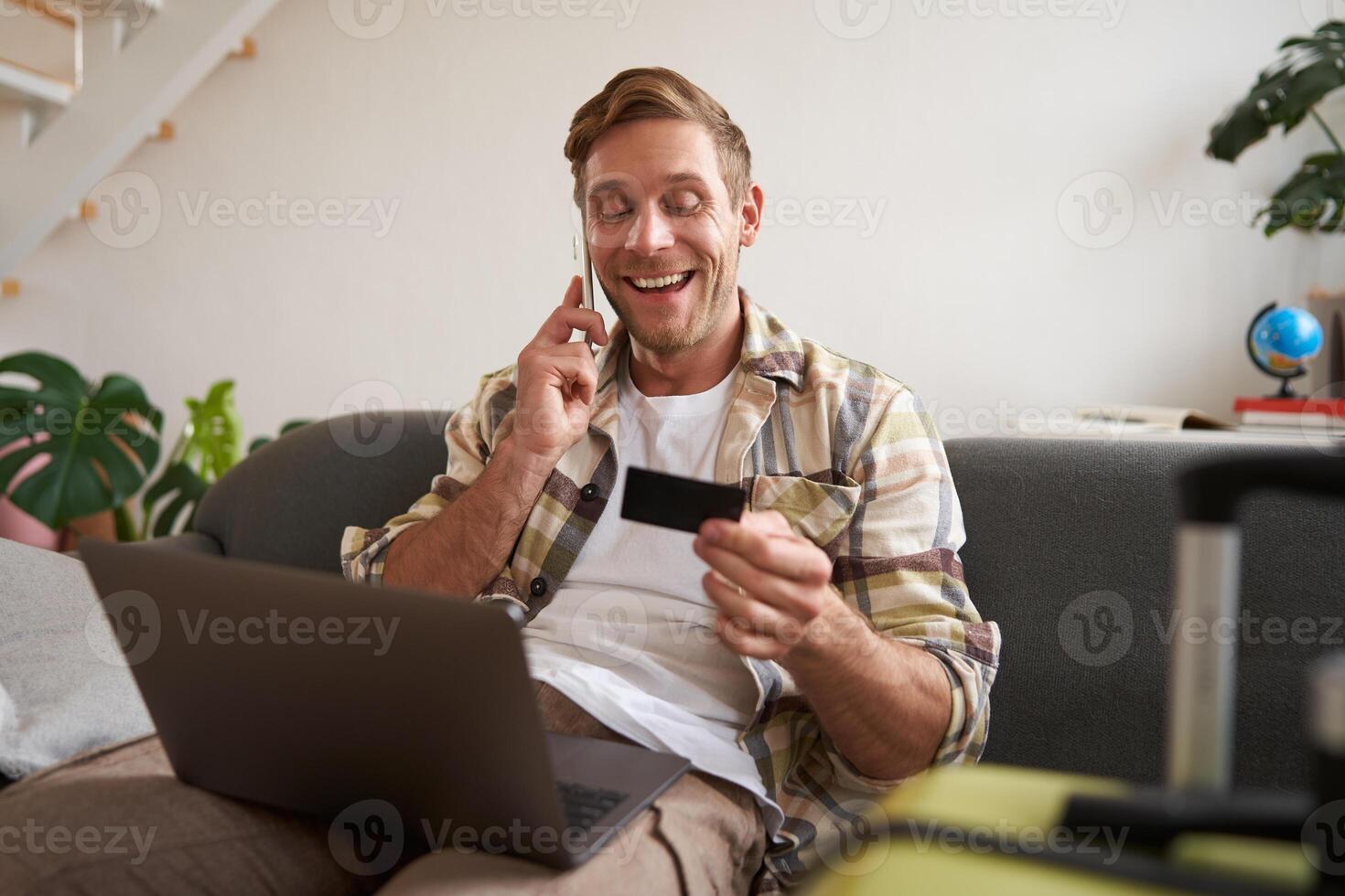 Portrait of young man with credit card, sitting and using laptop, making a phone call, booking hotel or tickets, confirming his purchase over telephone, has suitcase near him photo