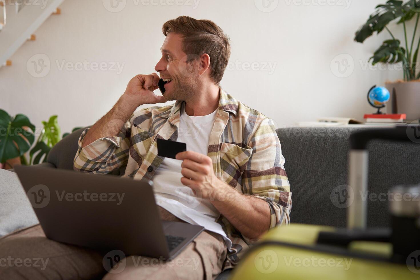 Image of guy calling someone, holding credit card and using laptop, has suitcase and camera packed for holiday photo