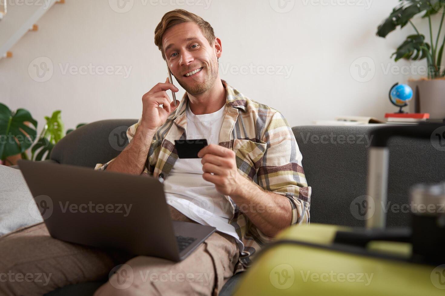 Portrait of young man with credit card, sitting and using laptop, making a phone call, booking hotel or tickets, confirming his purchase over telephone, has suitcase near him photo