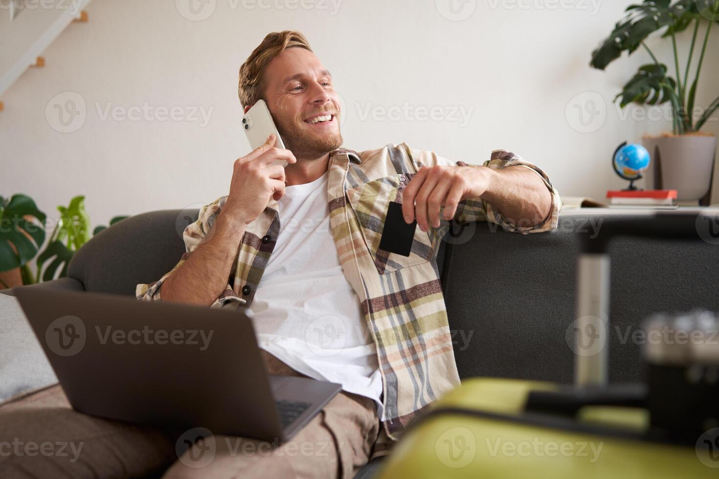 Smiling guy talking on mobile phone, sitting with laptop, holding credit card, looking out of the window with happy, relaxed face expression photo