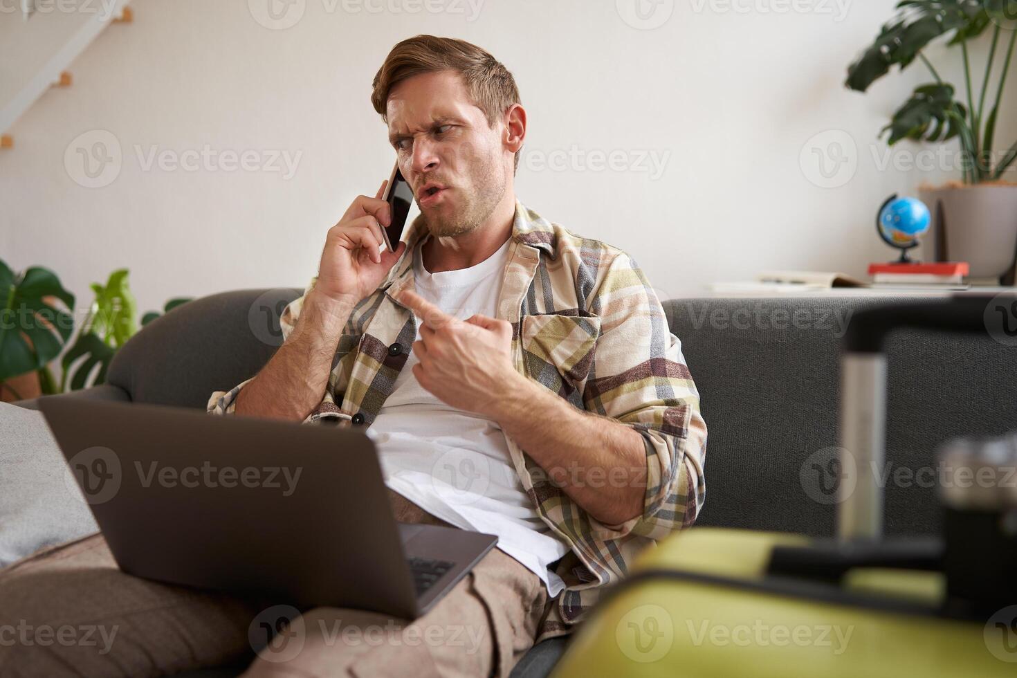 Portrait of angry man arguing over the phone, has laptop opened on his laptop, sits near suitcase, complaining at travel agency photo