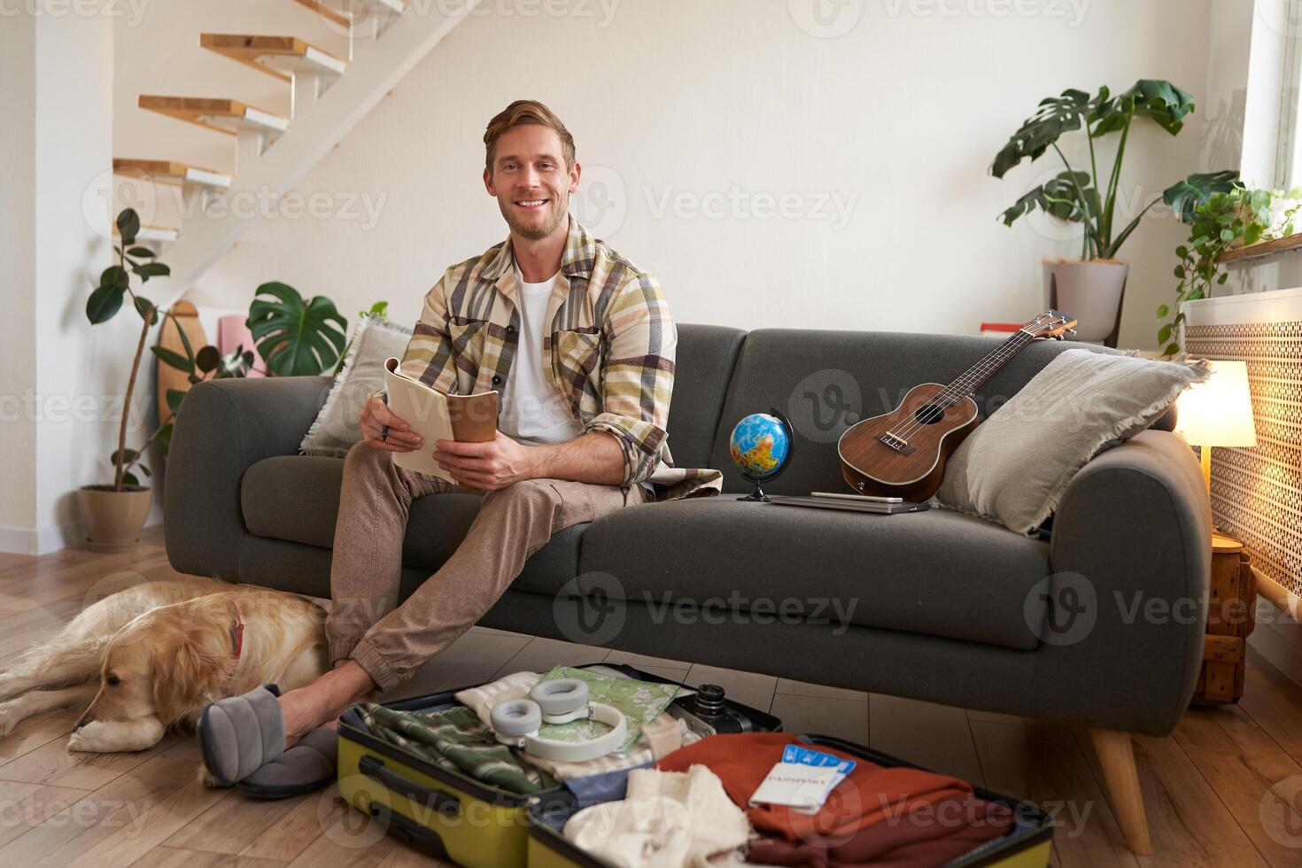 Portrait of young man with check list of items in hands, goes on holiday with his dog, packs suitcase for vacation photo