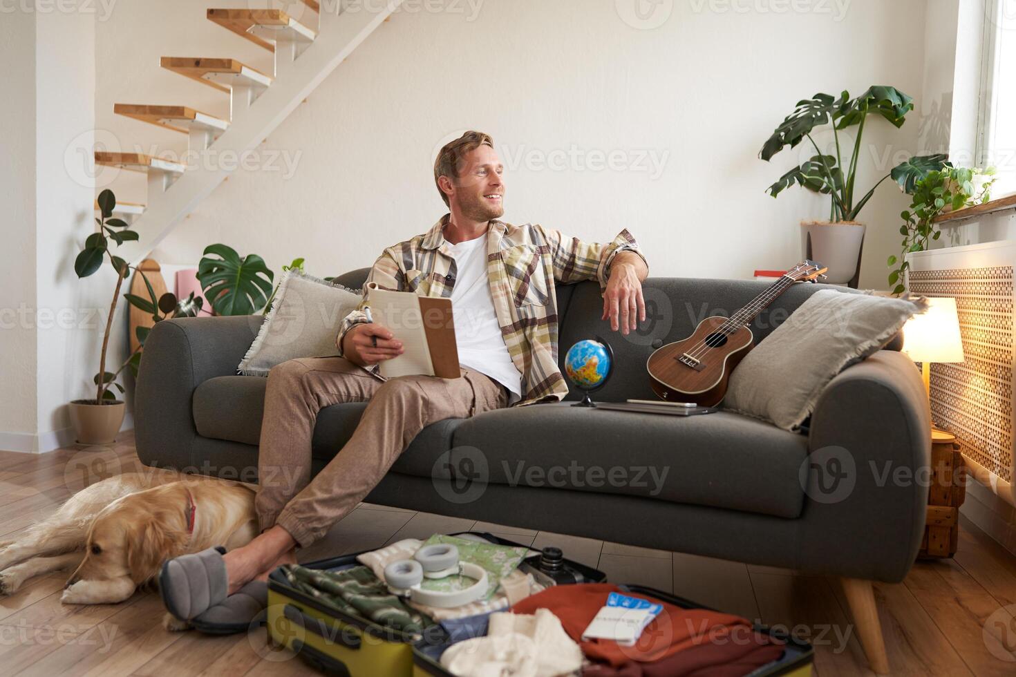 Portrait of young man sitting on sofa with notebook, has dog lying next to him, packing suitcase for summer vacation photo