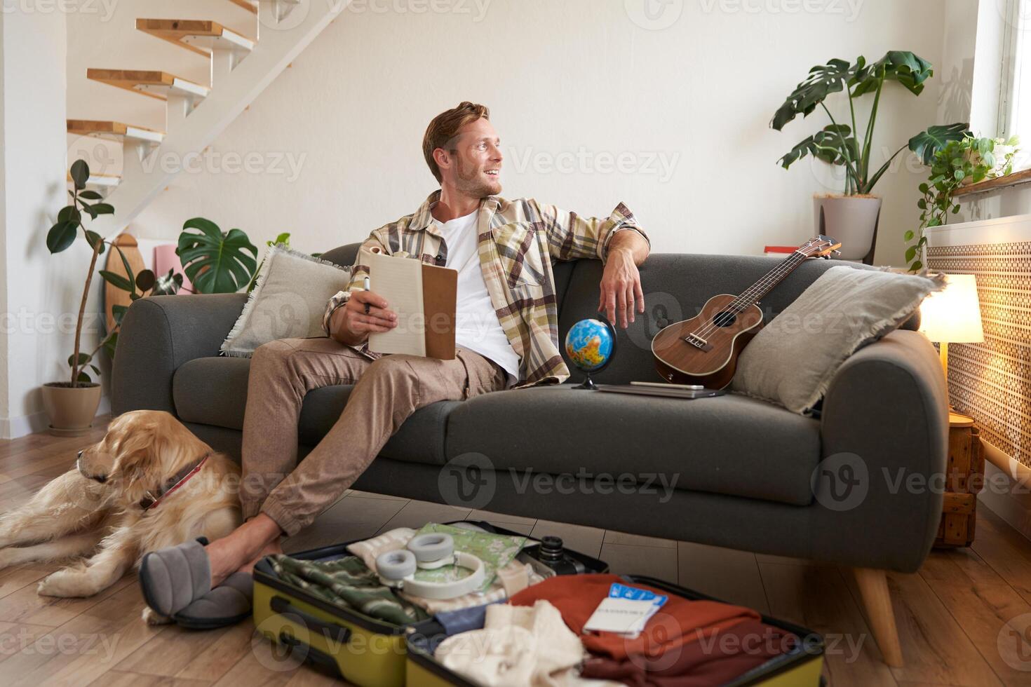Portrait of young man with check list of items in hands, goes on holiday with his dog, packs suitcase for vacation photo