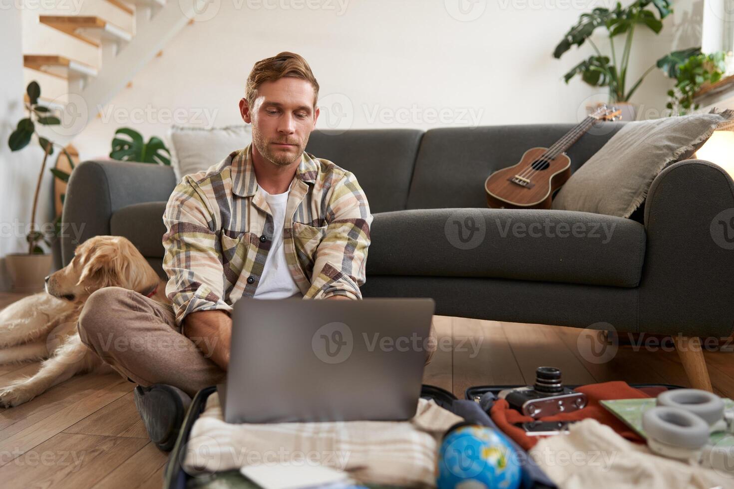 Man with laptop on suitcase, sits with dog on floor, looks online, browsing websites, packs clothes for vacation, goes on holiday, plans trip abroad photo