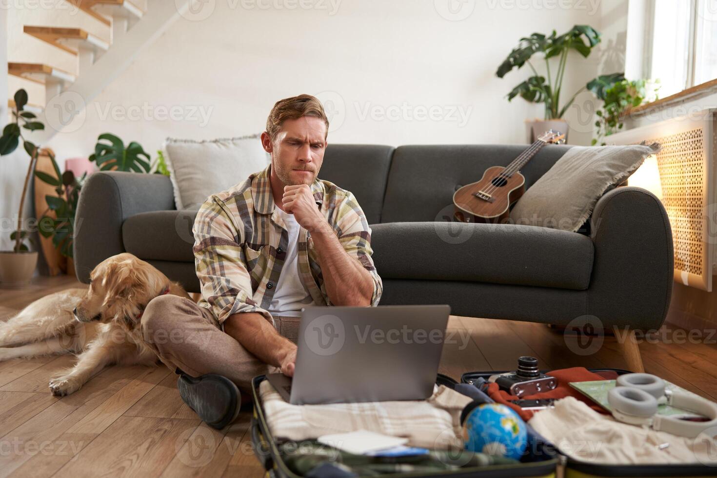 Image of young man, tourist going on holiday, sitting with a dog in living room, looking at laptop, check-in on a flight on computer photo
