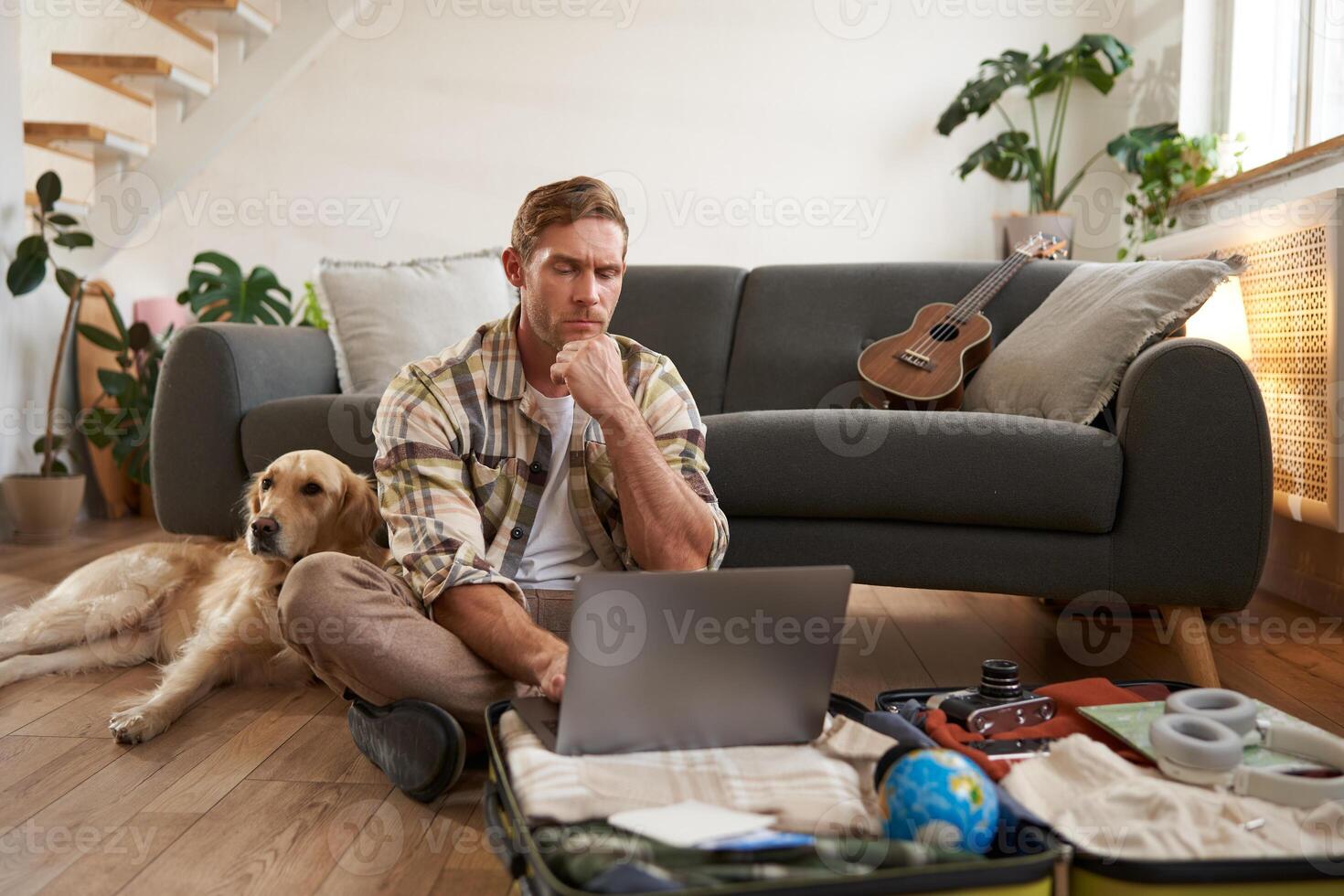 Image of young man, tourist going on holiday, sitting with a dog in living room, looking at laptop, check-in on a flight on computer photo