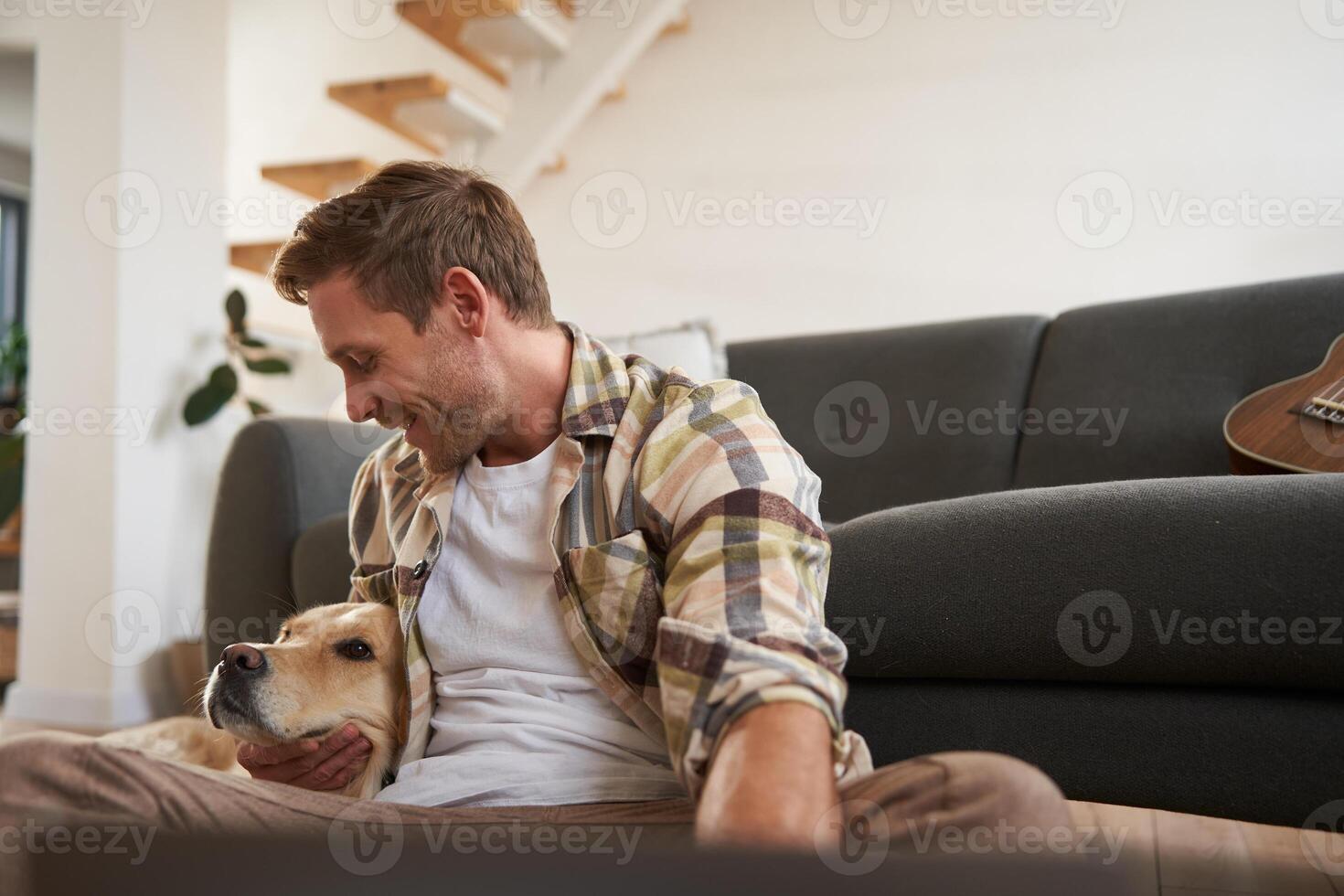 Portrait of handsome, happy young man, hugs his dog, playing with a pet, sitting on floor near sofa, using laptop photo