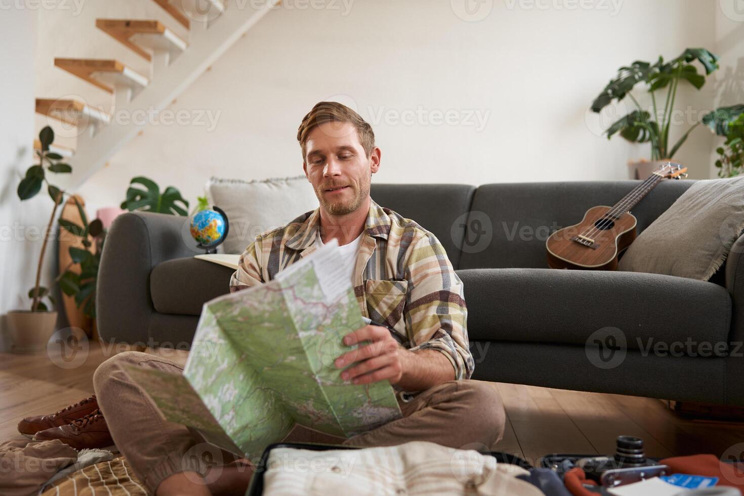 Portrait of happy young man, tourist with suitcase, sits in living room and looks at map, chooses holiday destination, picks route for travelling photo
