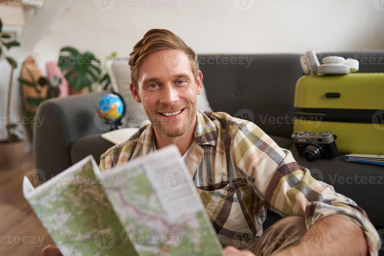 Portrait of handsome smiling man, holding map, sitting with suitcase in living room, going abroad on holiday, studying his route photo