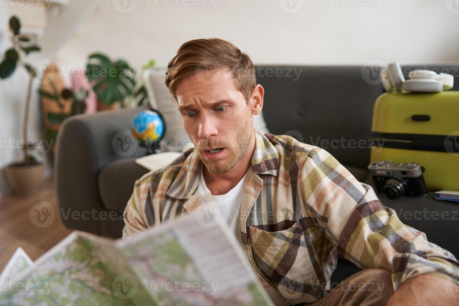 Portrait of man with concerned, puzzled face looking at map, frowning while studying the route, sitting with suitcase, going on vacation photo