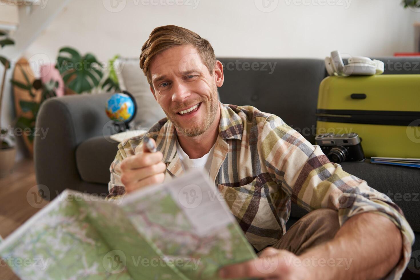 Happy young man with travel map, planning his journey, going on tour, sitting at home with suitcase photo