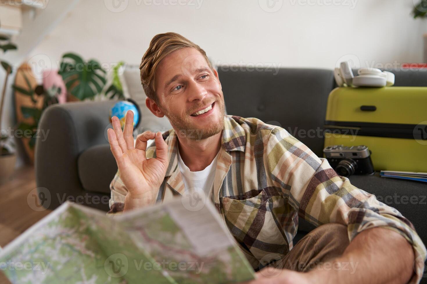Portrait of smiling handsome guy with travel map, plans tour, shows okay, ok hand gesture, sits on floor with packed suitcase photo