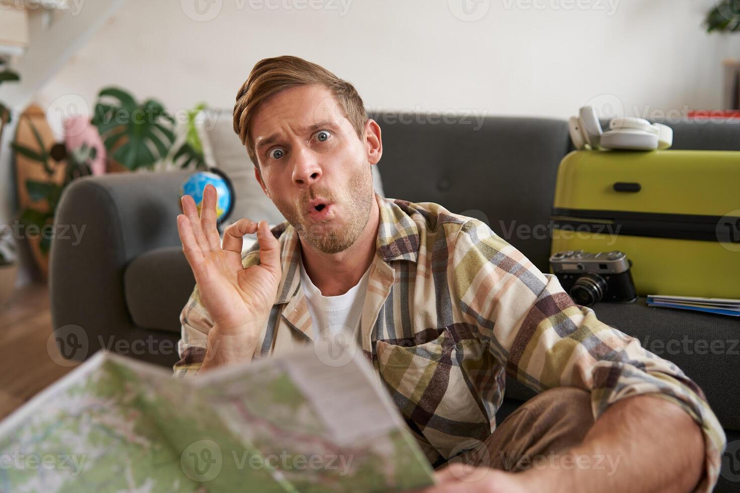 Portrait of smiling handsome guy with travel map, plans tour, shows okay, ok hand gesture, sits on floor with packed suitcase photo