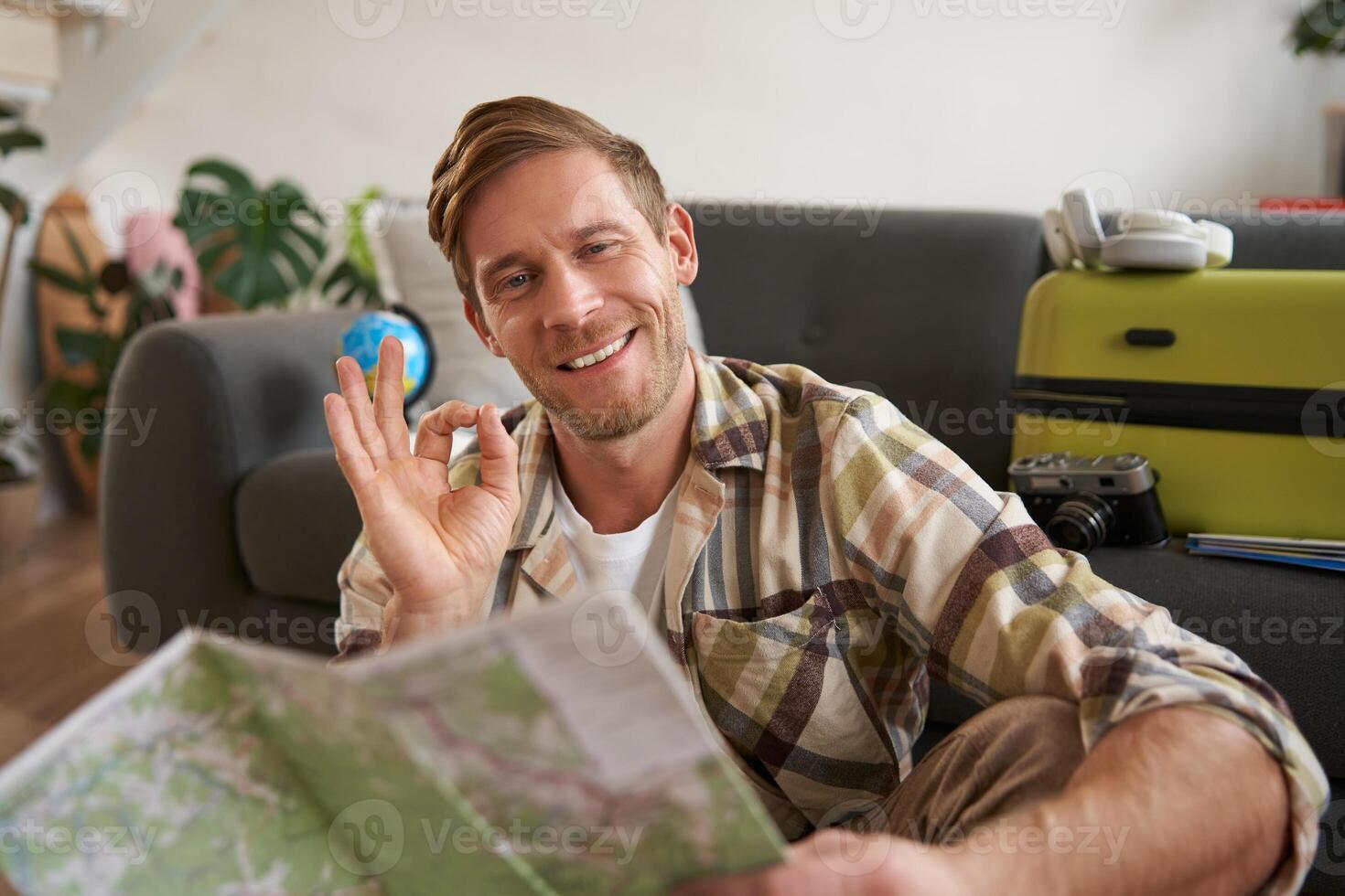 Smiling handsome tourist, man with road travel map, sits on floor near suitcase, shows okay, ok sign, recommending a tour photo
