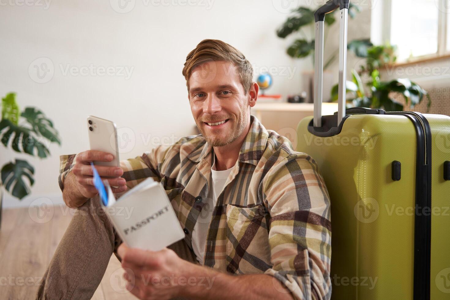 Portrait of smiling tourist, man sits with suitcase, holds smartphone, passport and tickets, goes on holiday photo