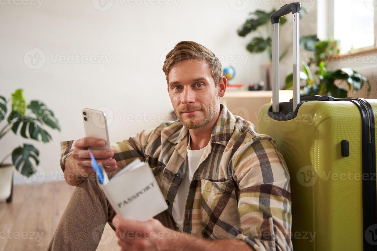 Portrait of handsome smiling man with suitcase, looks at smartphone, holds travel tickets and passport photo