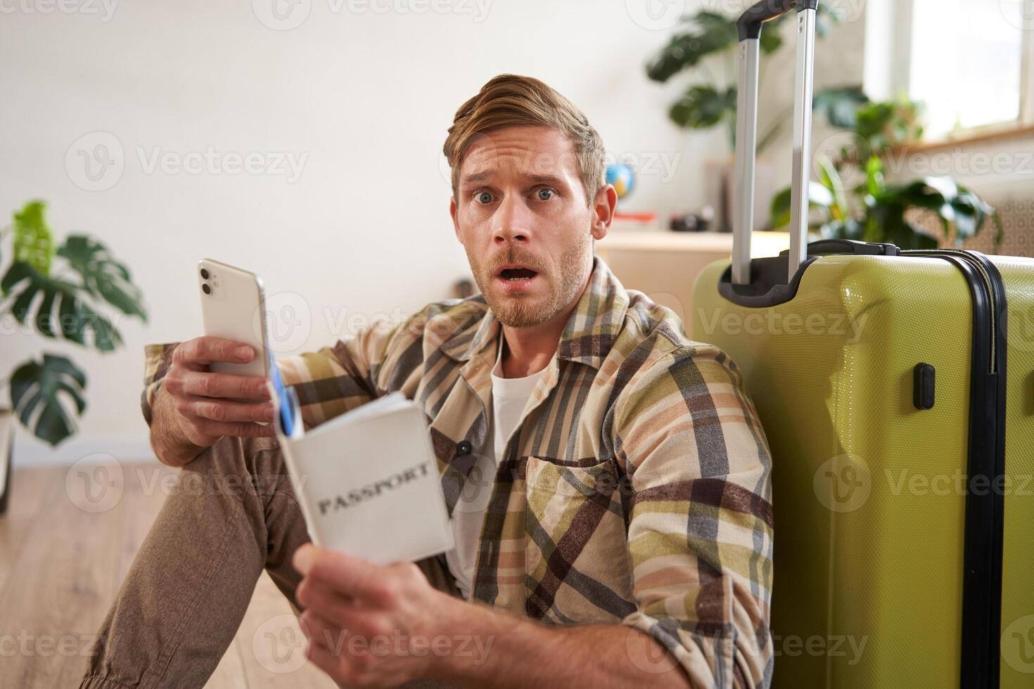 Man with scared face, sits on floor near suitcase, holds smartphone and passport photo