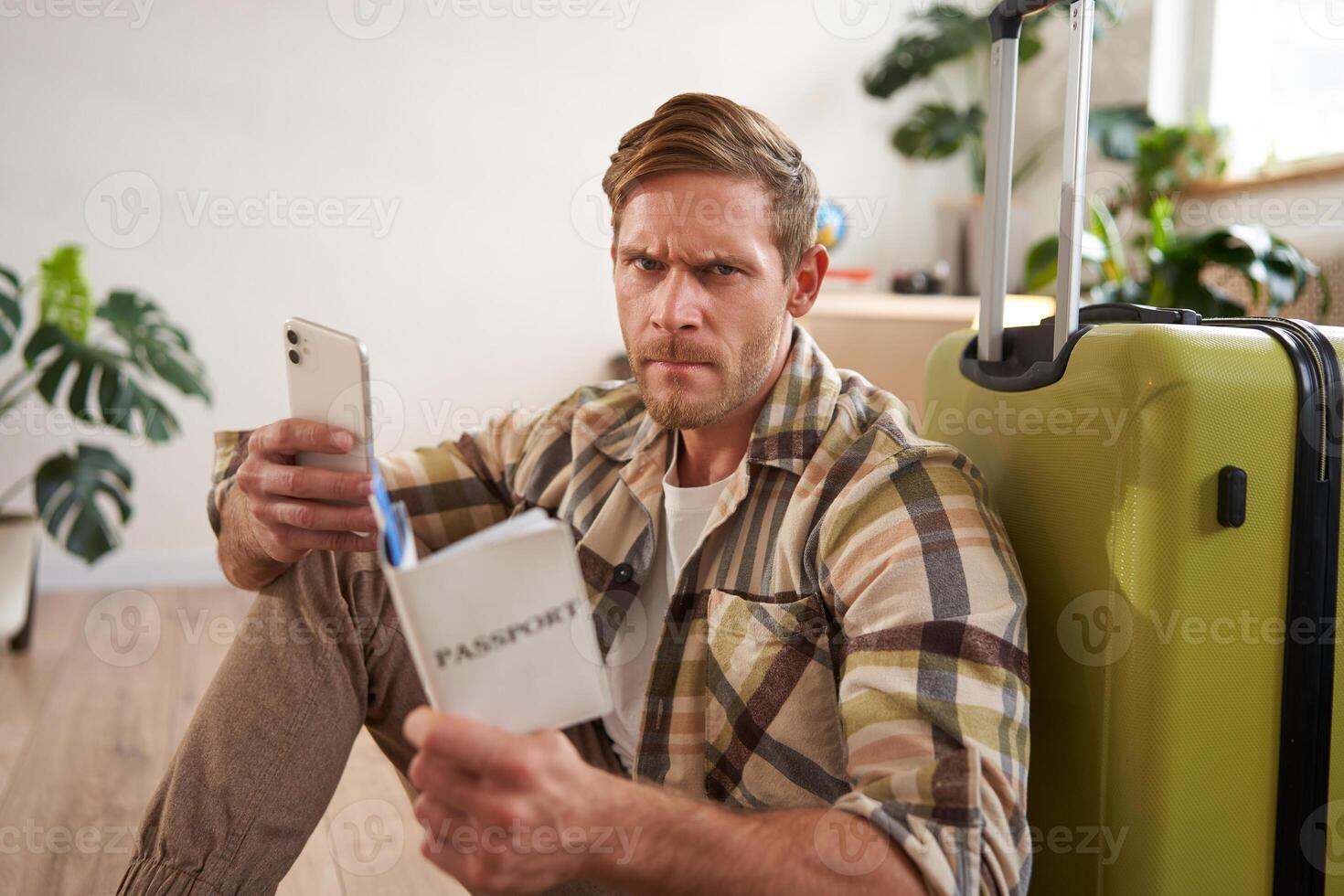 Angry man sits with suitcase, holds mobile phone and passport photo