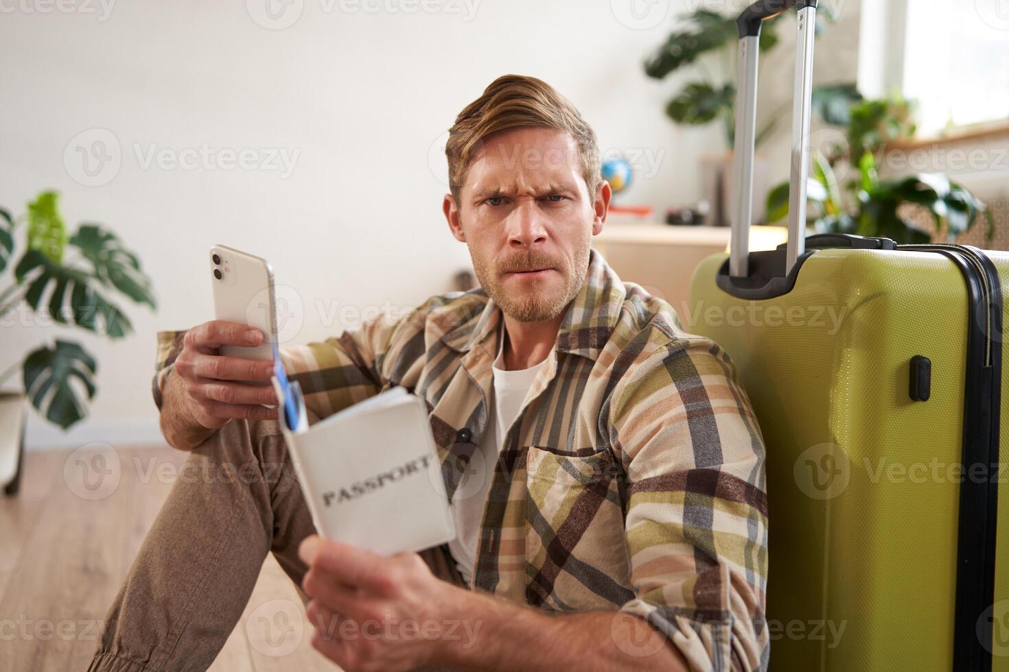 Portrait of angry man with suitcase and passport, holding smartphone, looking frustrated at camera, sitting on floor with luggage photo