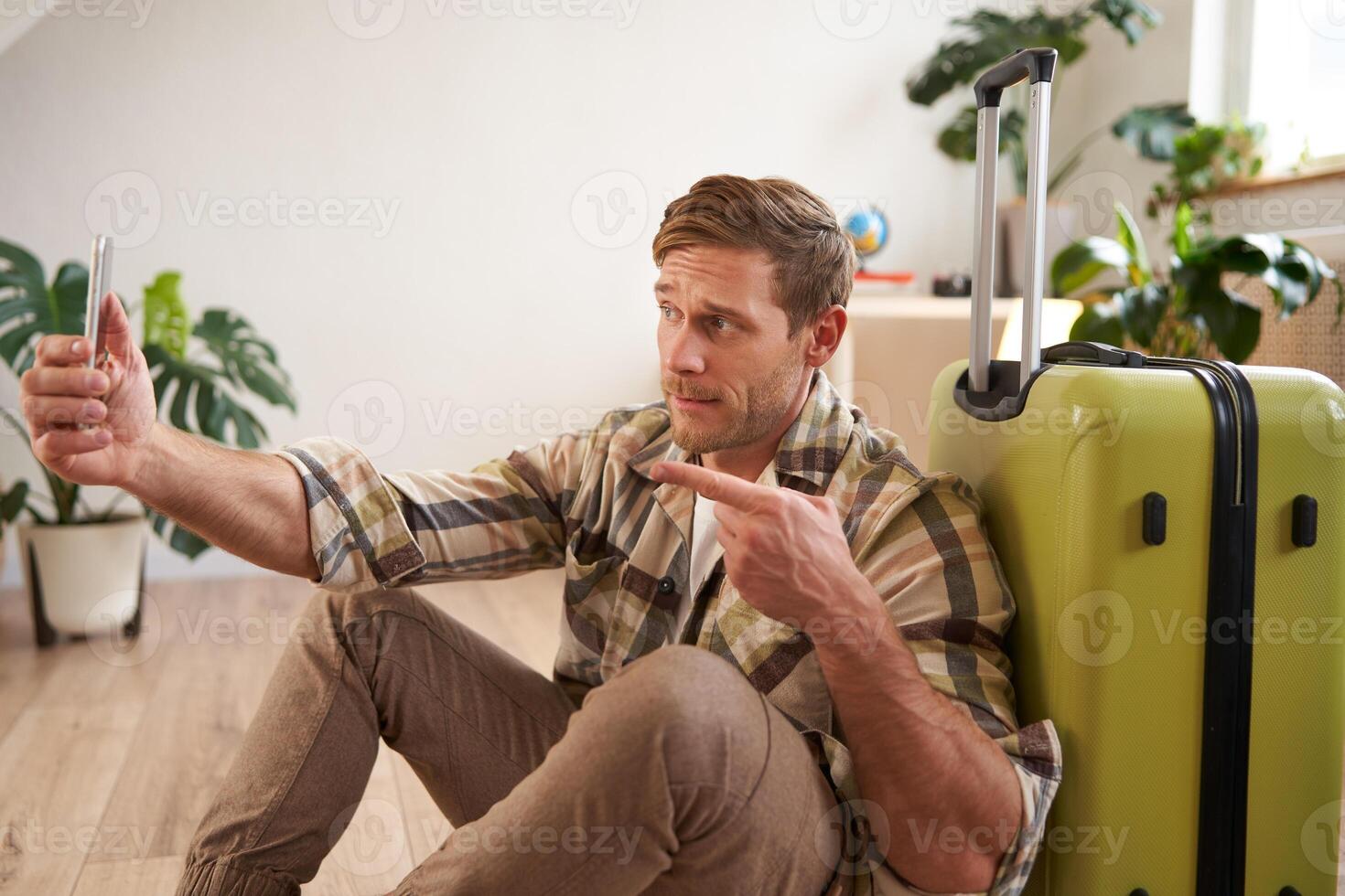 Portrait of handsome man goes on holiday, posts on social media about his vacation, going abroad, sits with suitcase, takes selfies on smartphone app photo