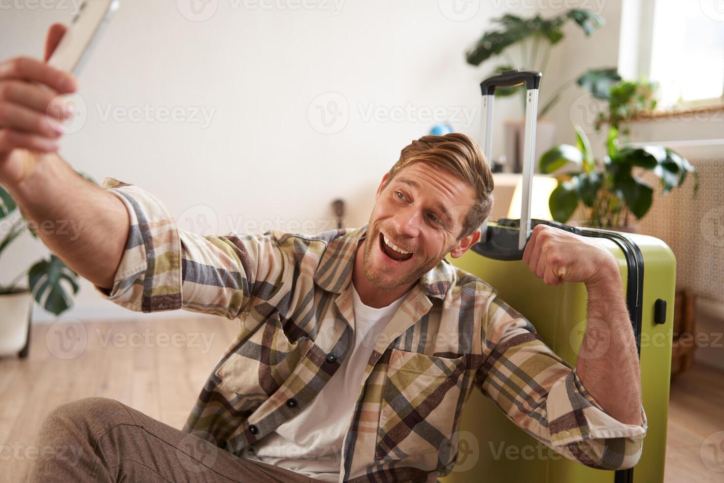 Portrait of smiling handsome guy shows his biceps on photo, takes selfies on smartphone, sits near suitcase, goes on holiday tour photo
