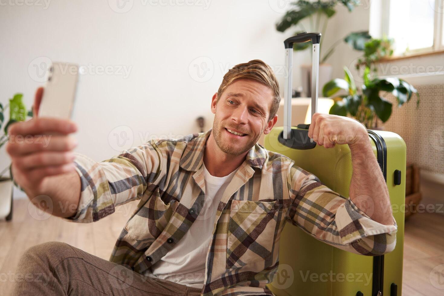 Image of handsome smiling young man going on holiday, posting online about vacation, taking selfie on smartphone with suitcase, shows muscles photo