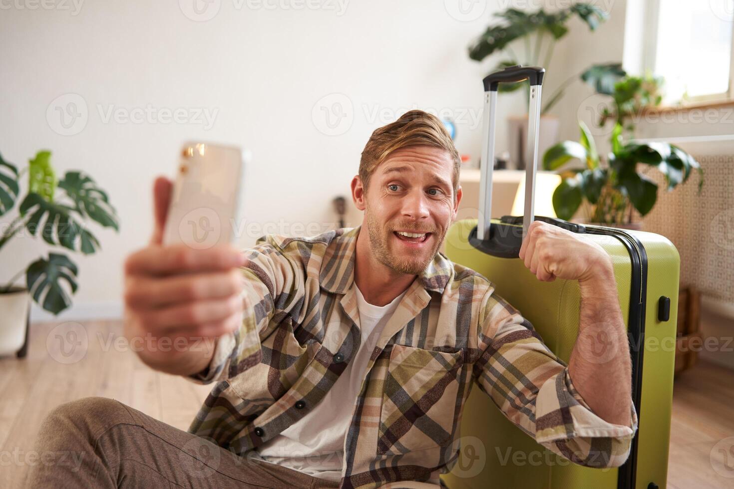 Funny handsome guy, tourist flexing biceps, goes on vacation, sits with suitcase, takes selfies on smartphone photo