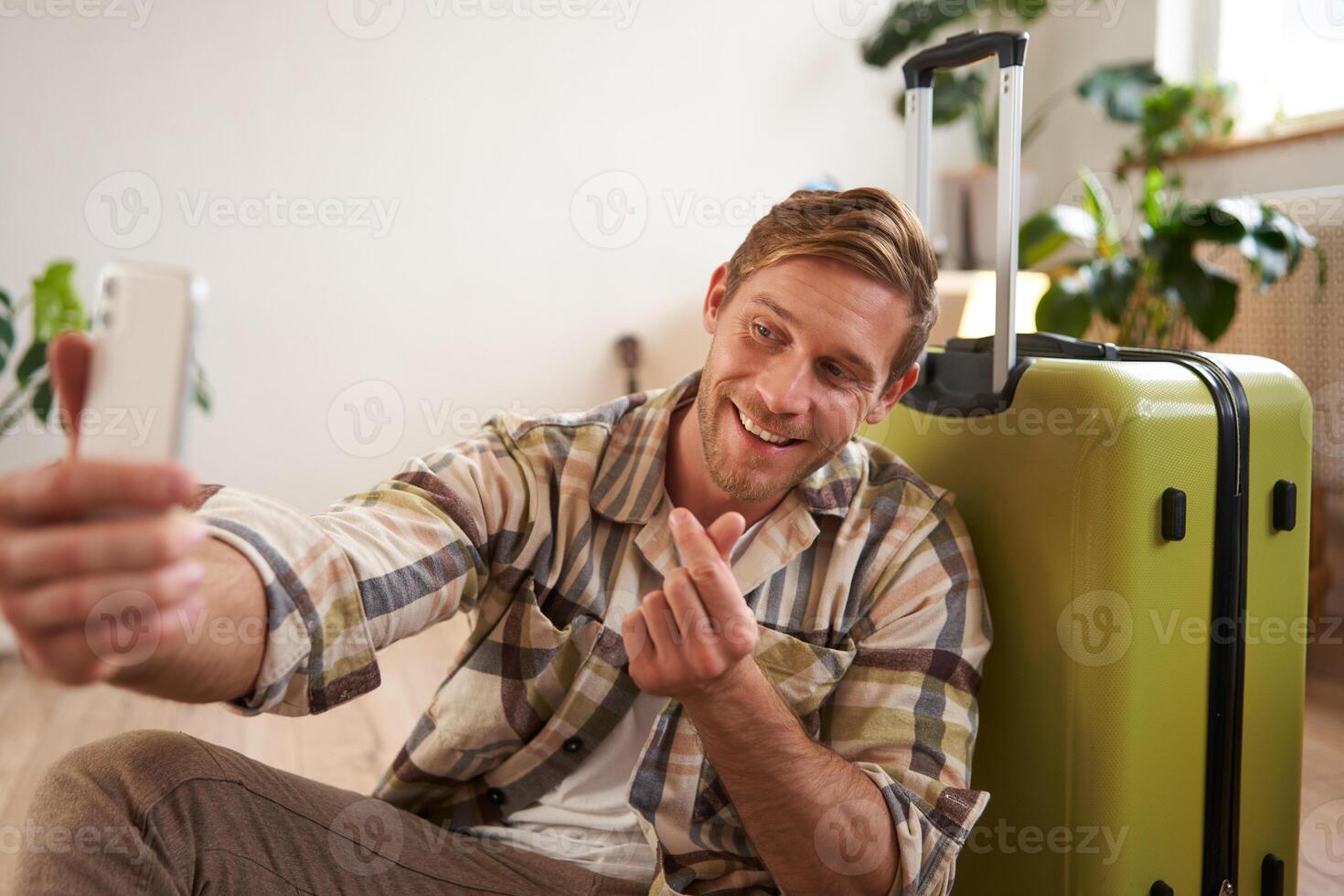 Portrait of smiling handsome guy, shows finger heart sign on selfie, takes photos of himself on mobile phone, poses with suitcase, goes on holiday