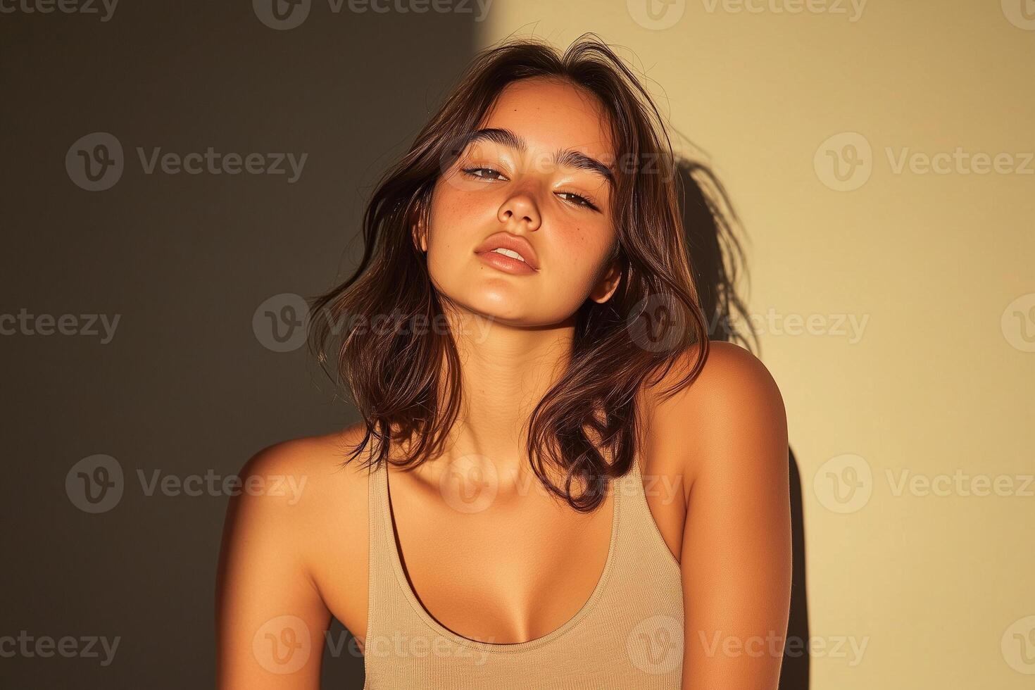A young woman with long brown hair and tanned skin poses in a beige tank top against a warm yellow background. photo