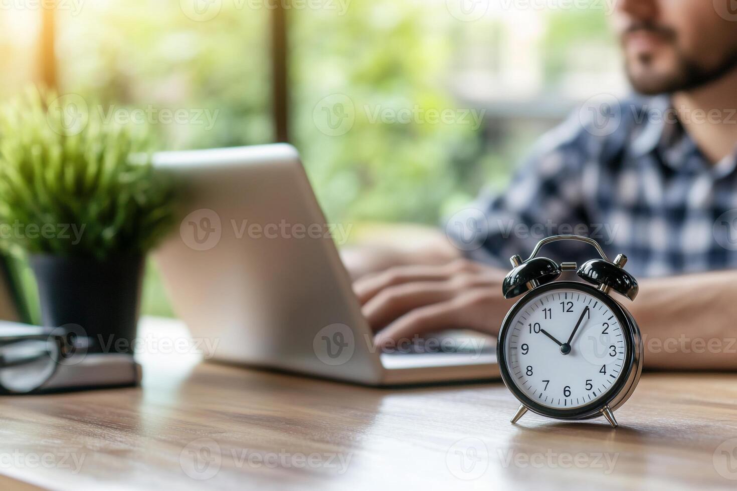 A person typing on a laptop with an alarm clock and a plant nearby. photo