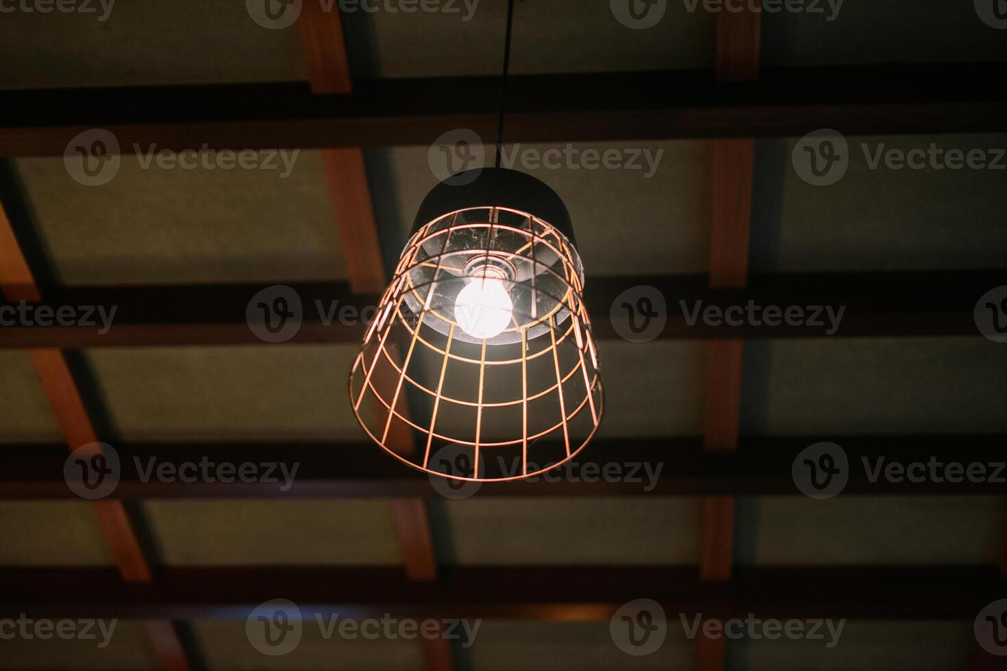 Illuminated caged bulb hanging from a wooden paneled ceiling photo