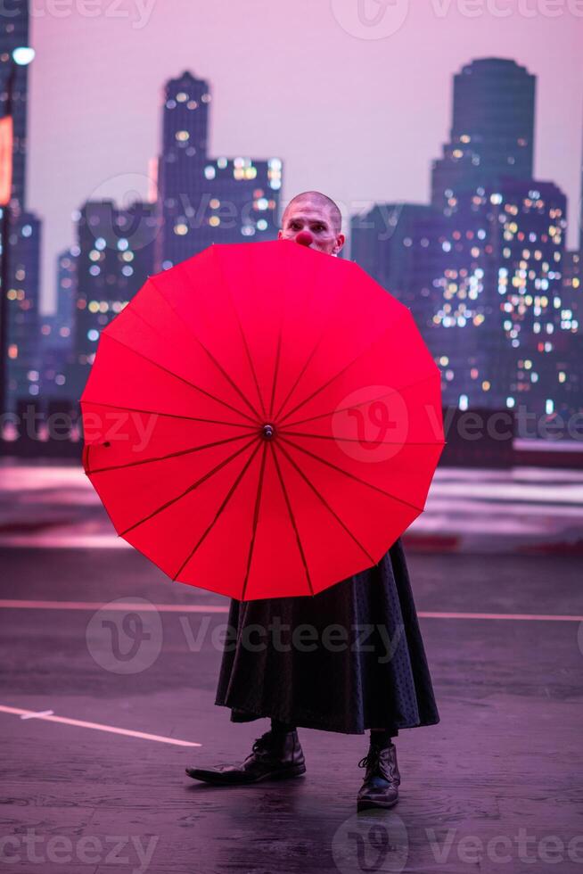 Mysterious Clown Performer with Red Umbrella in Urban Nightscape. photo