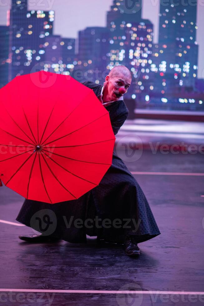 Mysterious Clown Performer with Red Umbrella in Urban Nightscape. photo
