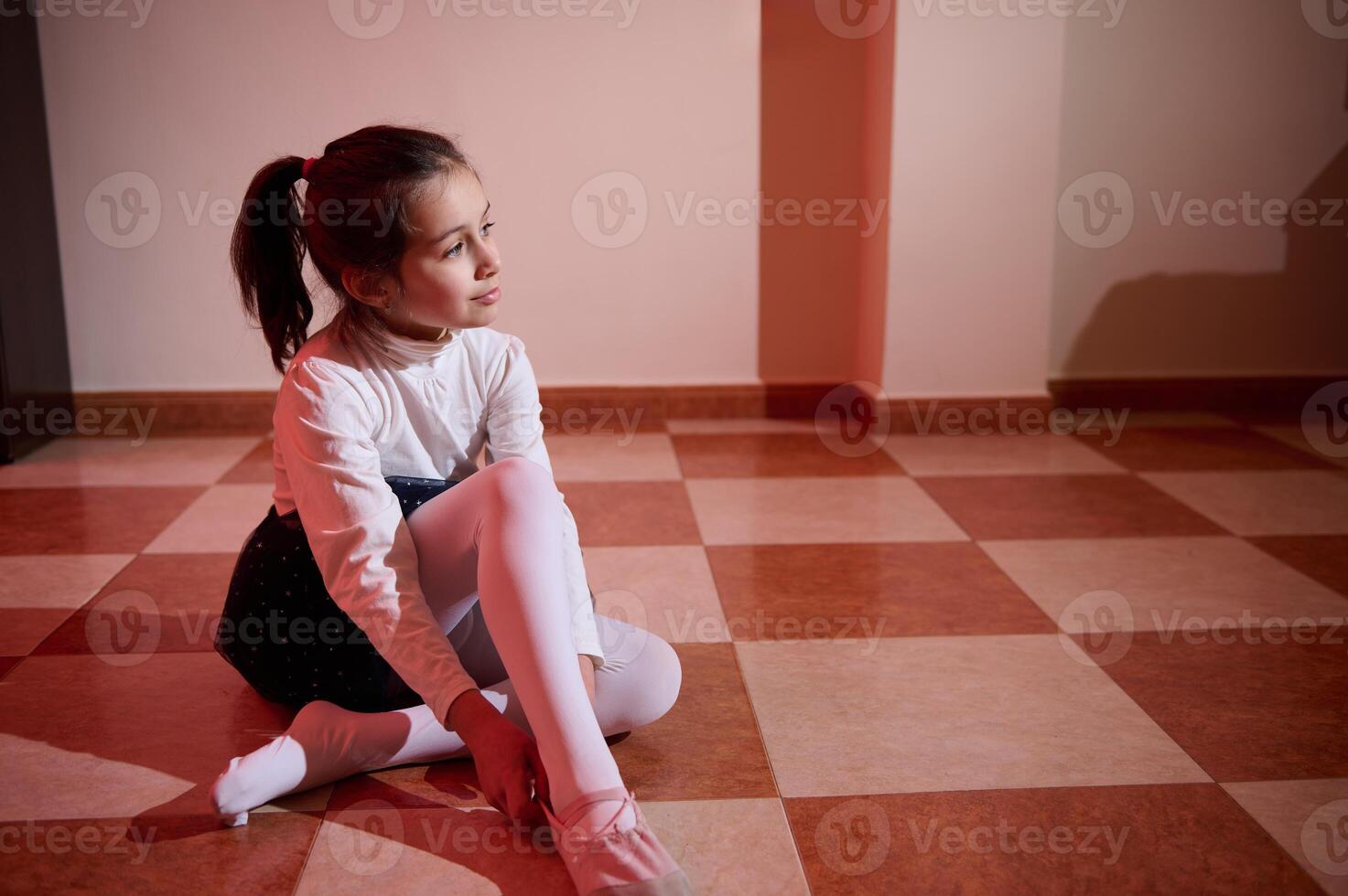 Young Girl Sitting on Tiled Floor in Ballet Attire Under Warm Lighting photo