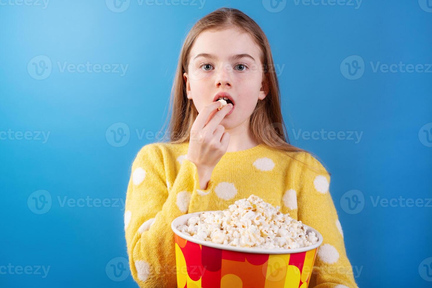 Girl eating popcorn from a bucket, wearing a yellow sweater, enjoying snacks with a surprised expression on a blue background. photo