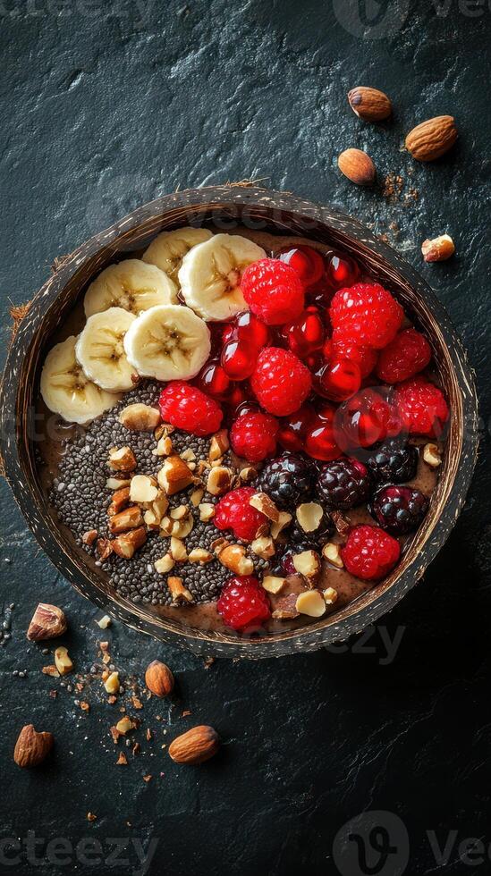 Healthy smoothie bowl topped with bananas, raspberries, blackberries, and nuts served on a black textured surface photo