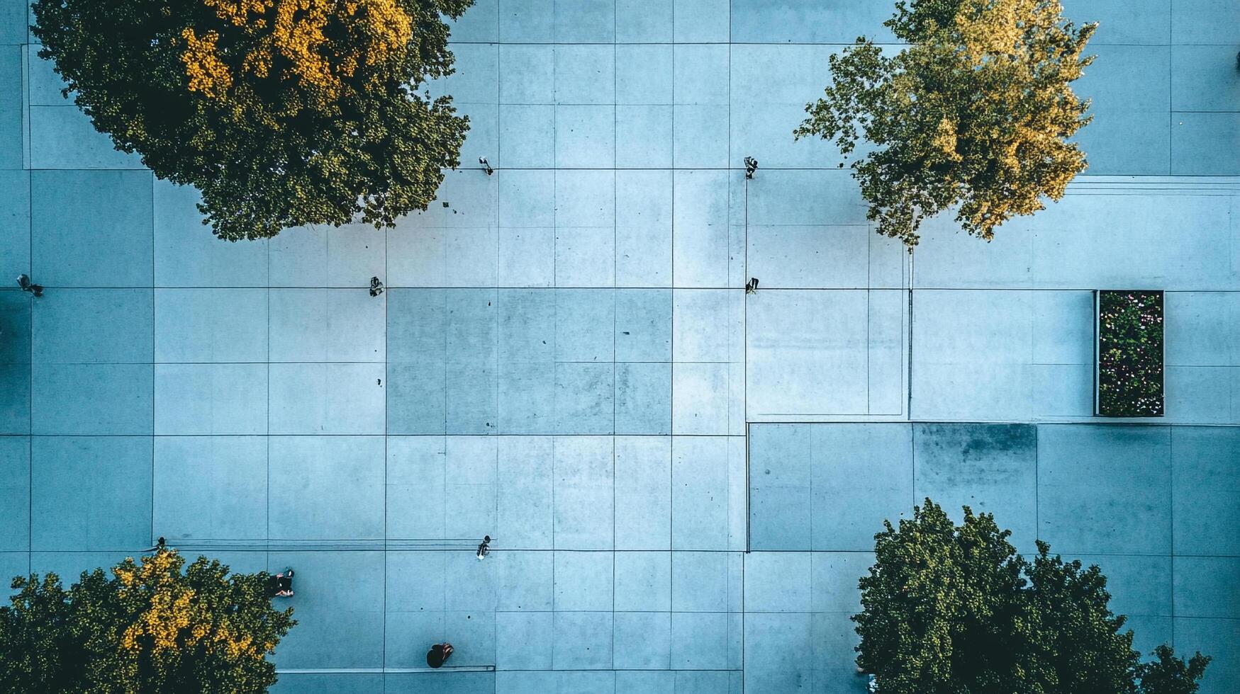 Aerial view of a city square. A concrete surface is populated by trees and a small green space, with people strolling around the area. photo