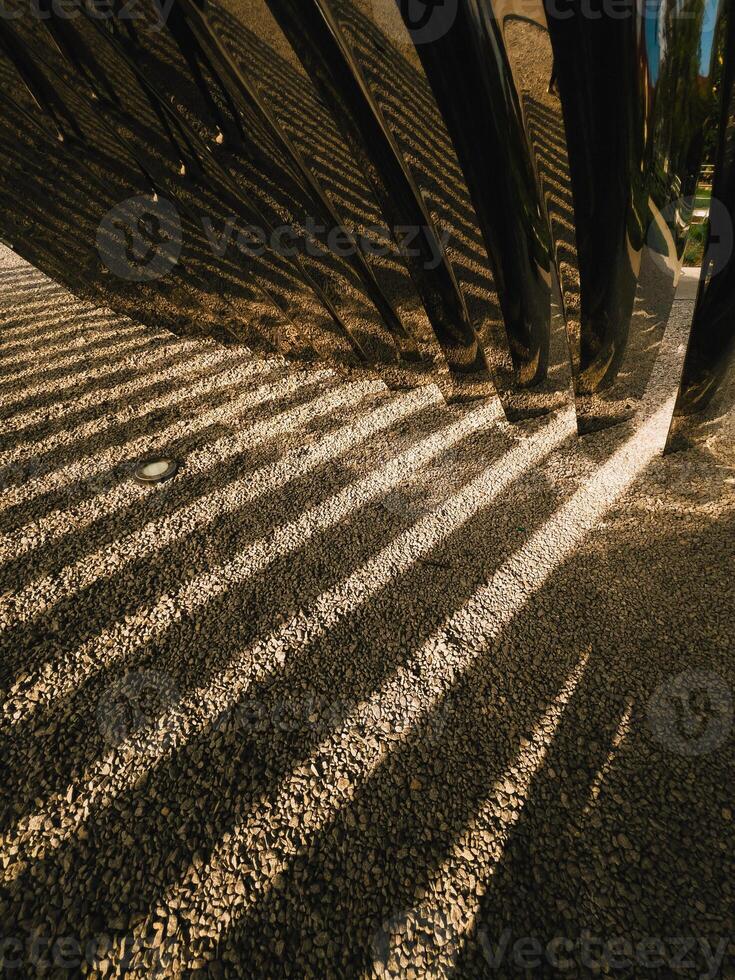Sunlight reflecting off polished metal tubes casts intricate shadows onto a gravel surface, forming abstract pattern of contrasting light and dark lines, enhancing the modern design. geometric shadows photo