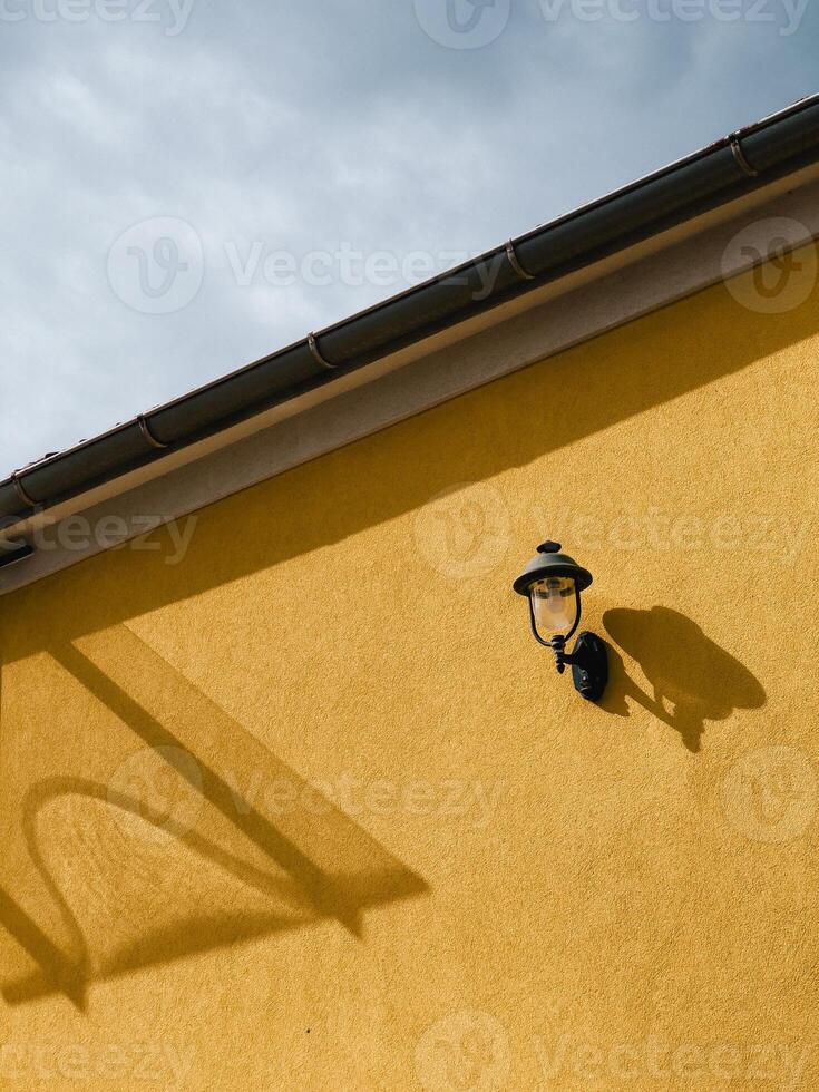 Outdoor lamp casts a shadow on a vibrant yellow wall, complemented by a gutter and a cloudy sky, creating a striking contrast of light and shadow in an urban setting. Lamp and bright yellow wall photo