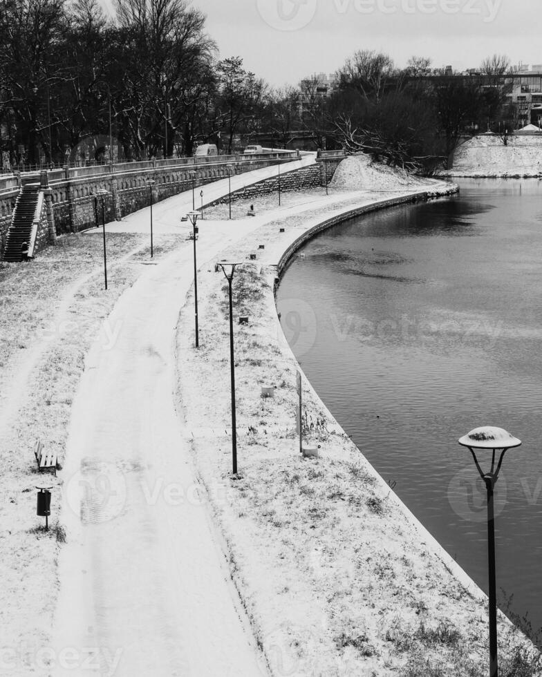 Snow covered promenade lining the river in winter, featuring street lamps, benches, and trash cans, captured in striking black and white image that evokes tranquility and solitude. promenade in winter photo