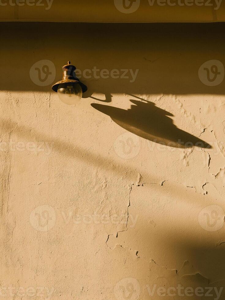 Vintage lamp casting long shadows across weathered wall, golden sunlight illuminating rustic surface during warm late afternoon light photo