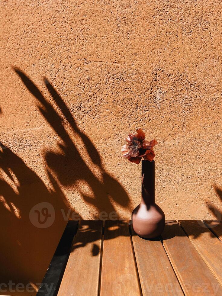 Dried botanical resting in earthy ceramic vessel. Minimalist composition featuring a dried flower in a brown ceramic vase on a wooden table, casting dramatic shadows against a textured orange wall photo