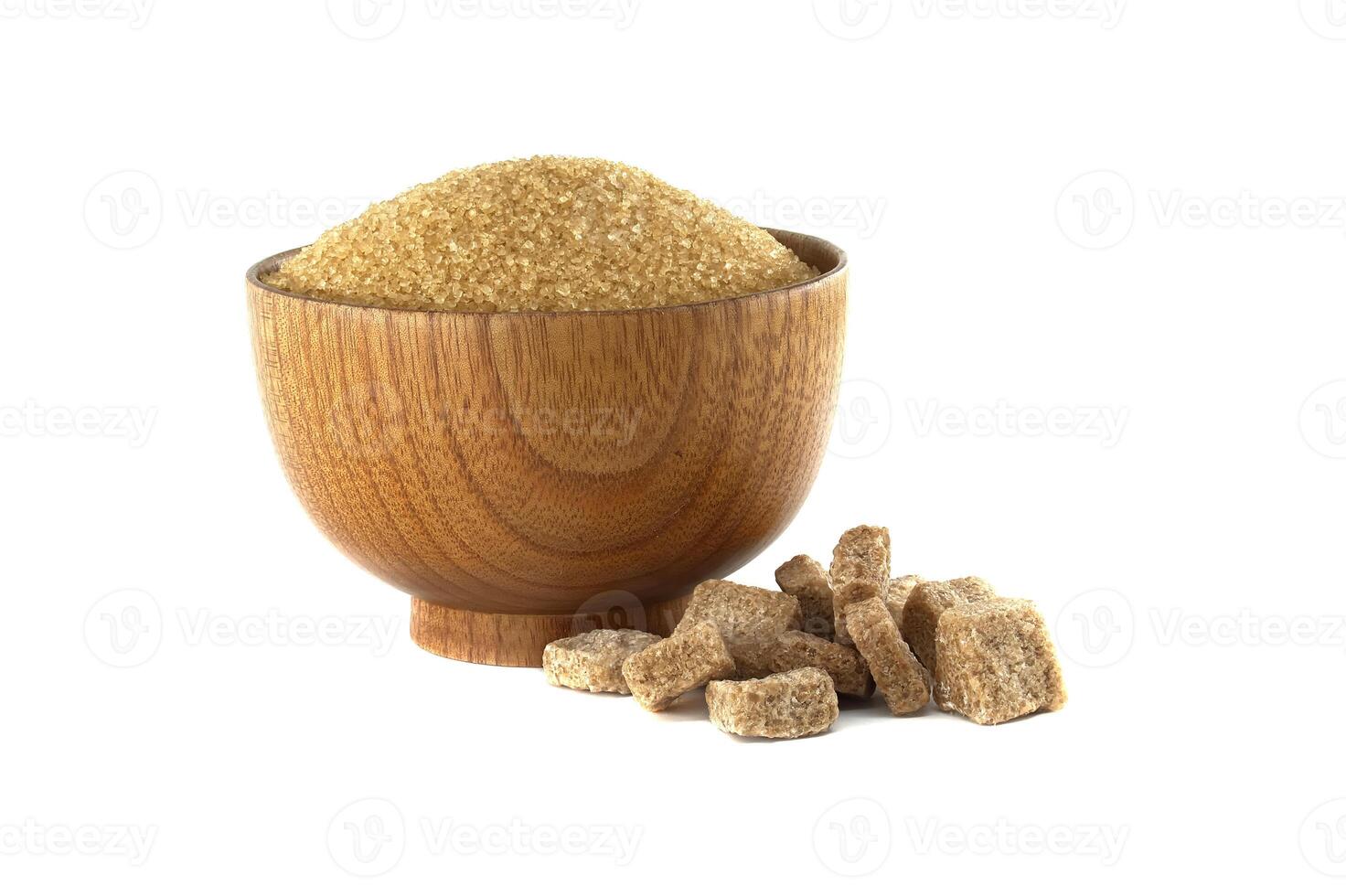 A wooden bowl overflows with raw brown sugar granules, complemented by scattered sugar cubes. photo