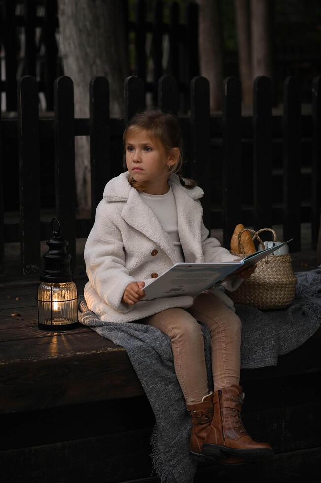 Under the soft lantern light, a girl enjoys her book while sitting outside, a basket filled with fresh bread resting nearby. photo