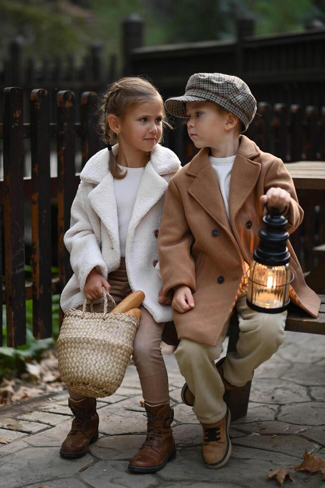 young boy and girl sit closely on a rustic bench dressed in timeless vintage attire. the warm glow of their lantern contrasts and basket beside them adds charm of their nostalgic moment photo
