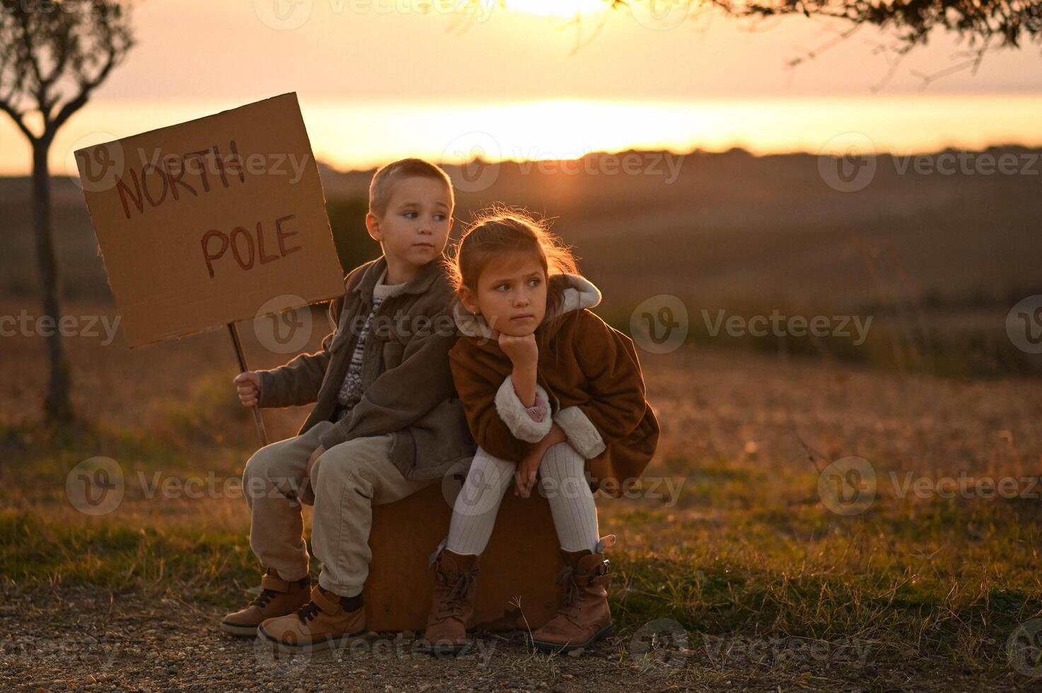 Dressed in vintage outfits, the kids sit on a worn suitcase, bathed in golden light. They gaze into the distance, hoping for someone to take them to the North Pole. photo