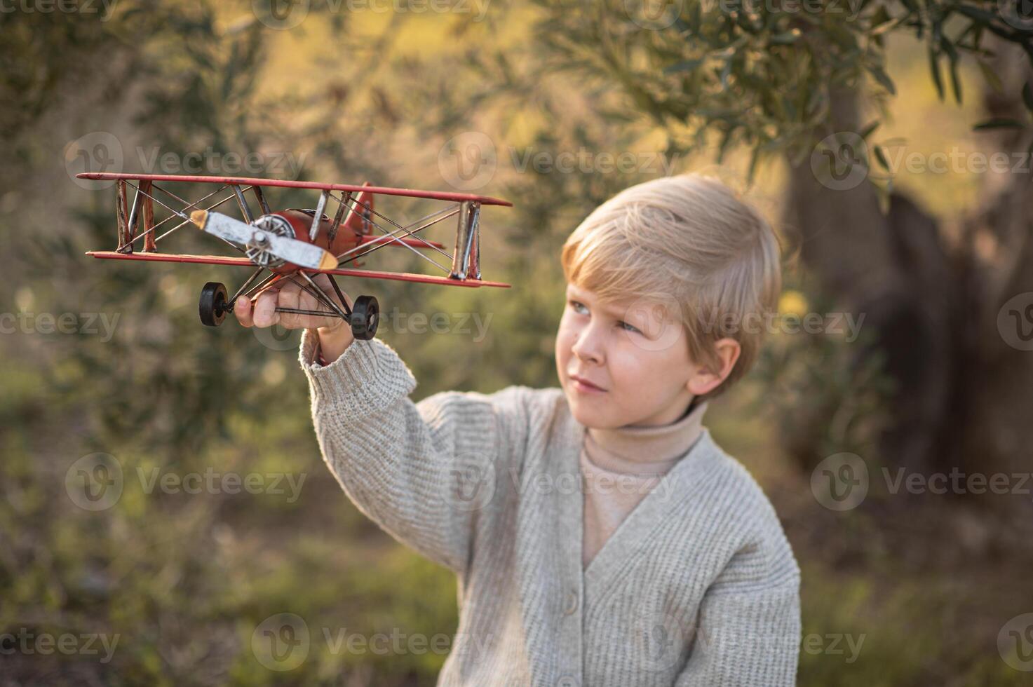 A young boy sits on an old suitcase under a tree, playing with a vintage airplane photo