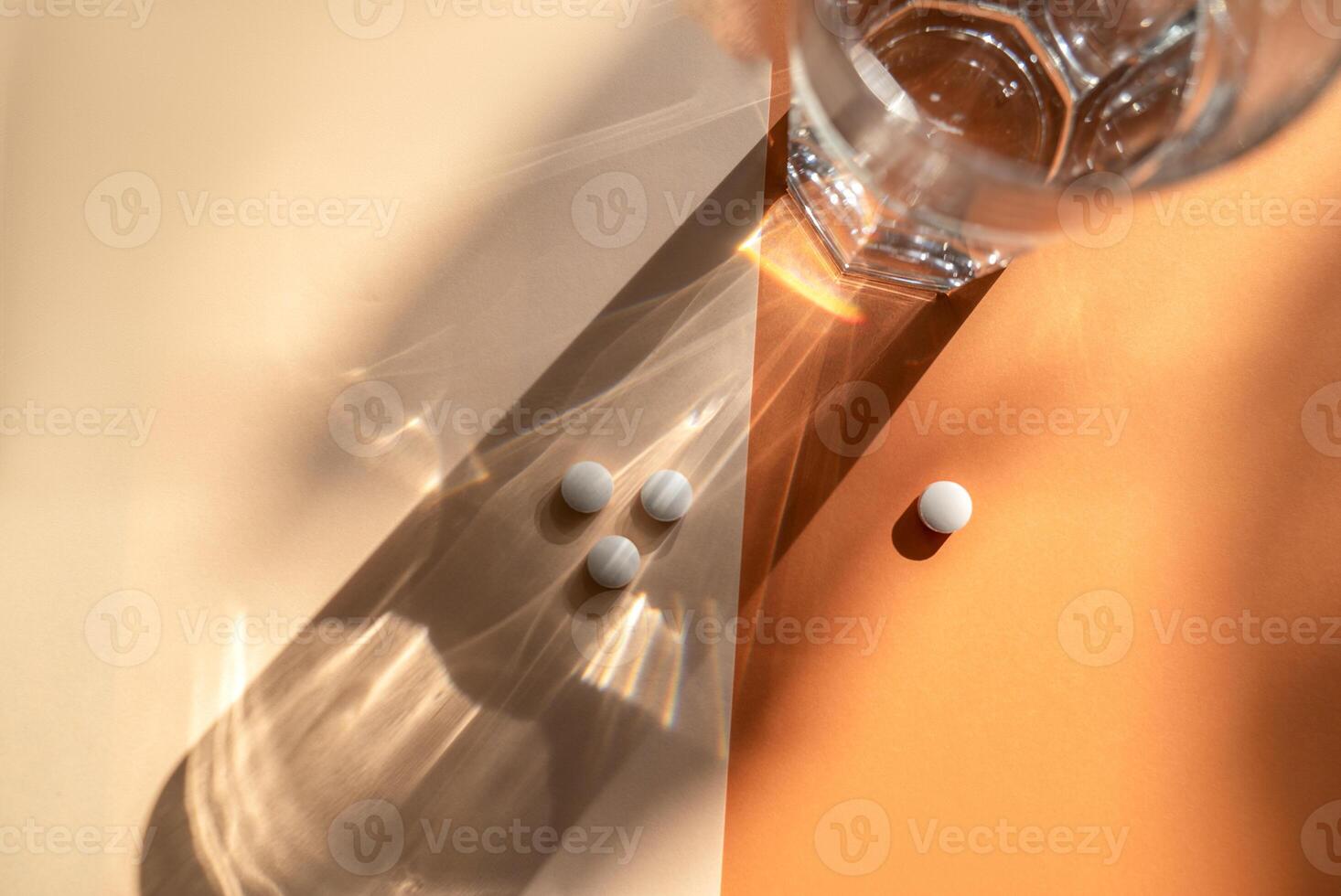 White medical pills lie on the background, next to a transparent glass of water. A shadow from a glass of water falls on an orange background. Medical concept of taking medication. White, round pills. photo