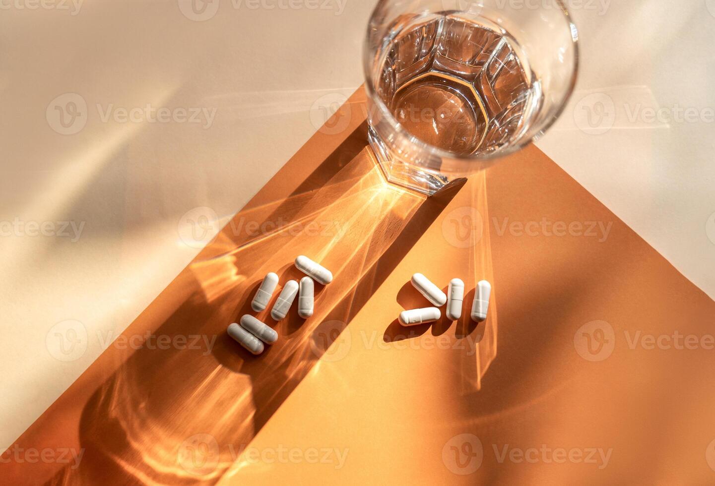 white medical capsules lie on the background, next to a transparent glass of water. A shadow from a glass of water falls on an orange background. top view. medical concept about medicine. two groups of capsules photo